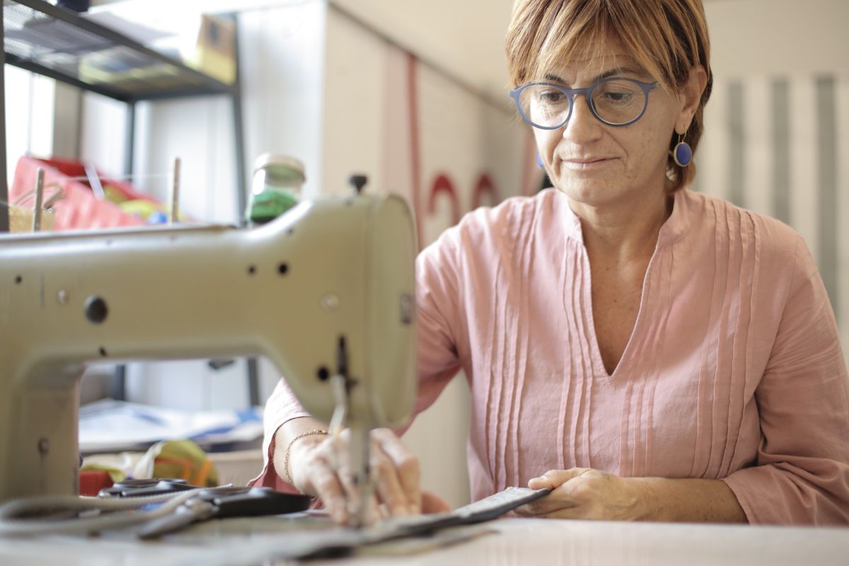 A mature seamstress guides fabric through a sewing machine, illustrating continued work while considering Medicare and group health insurance.